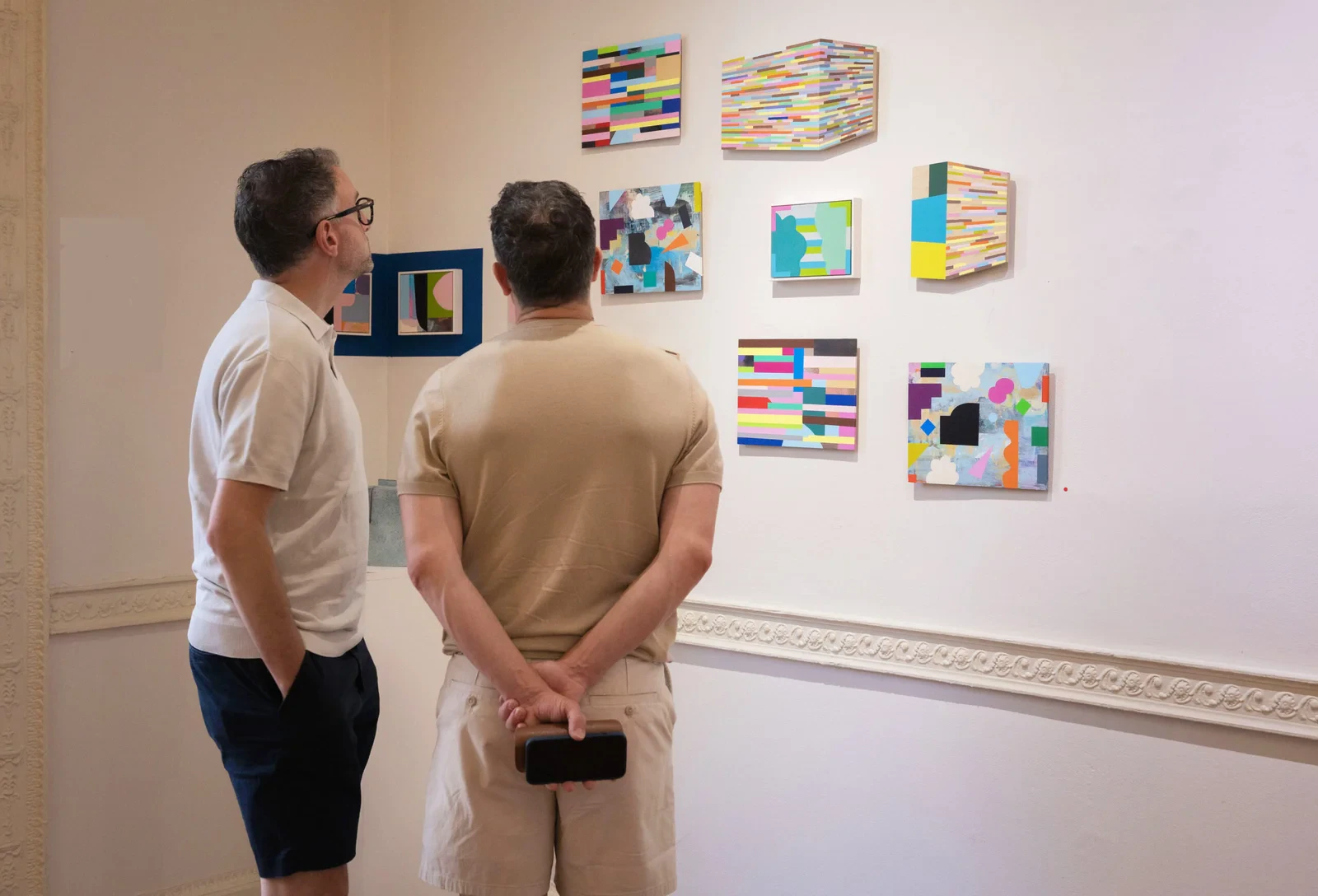 Two visitors viewing a wall of Mary O'Connor's small abstract geometric paintings at an exhibition