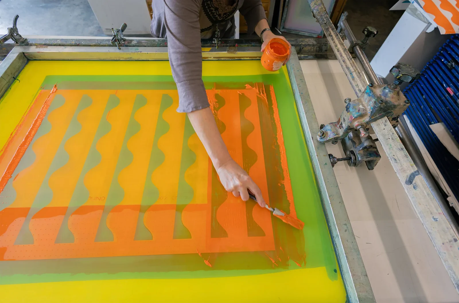 Mary O'Connor hand-pulling a screen print — orange ink drawn across a yellow-green screen in the studio